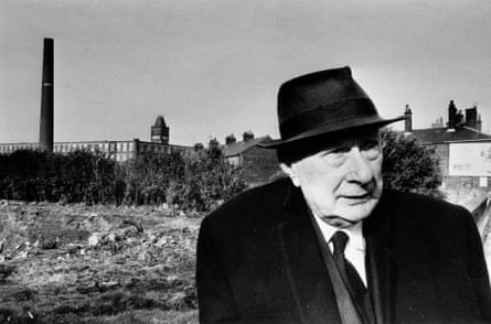 LS Lowry in a hat, coat and tie with a mill in the background