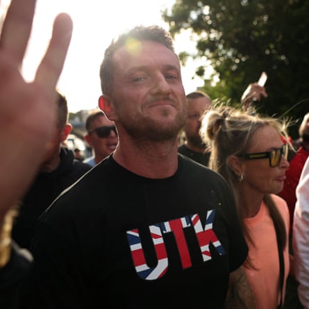 Man with short hair in T-shirt featuring red, white and blue lettering, smirks into lens.