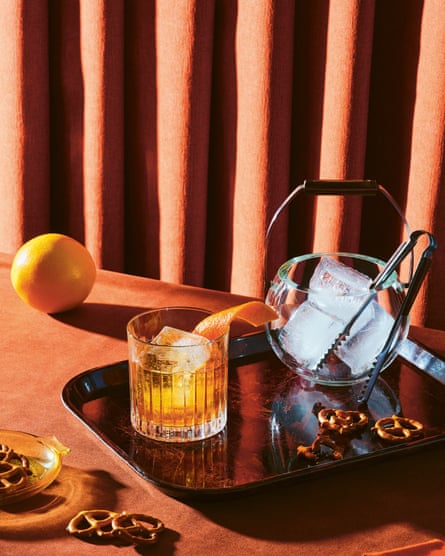 A old fashioned cocktail on a tray, next to a bowl of ice. The tray is decorated with pretzels
