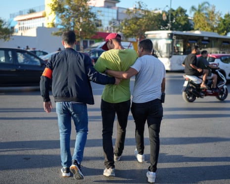A young Arab man seen from behind is frogmarched across a main road by two men in civilian clothes