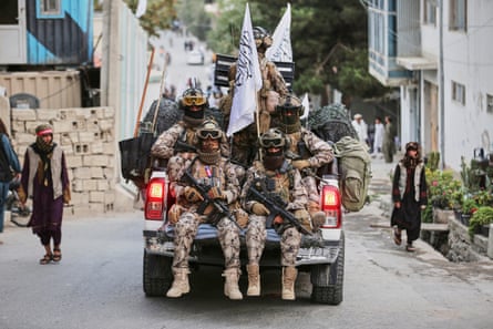 Taliban fighters in desert camouflage fatigues, masks and big boots sit in the back of a pickup truck; they carry machine guns and white flags with Arabic script; the truck is going down what looks like a residential street with flowers growing in front of a building. Two men are walking up the hill on either side of the street, both in traditional loose clothing wearing sandals and with scarves over their heads