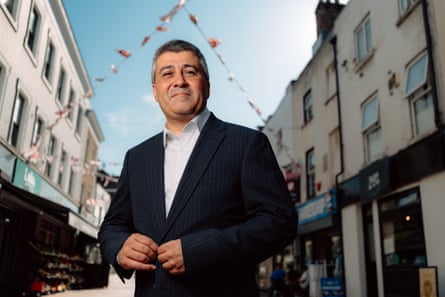 Sunder Katwala stands smiling in the middle of a high street of shops in Dartford, Kent. There are flags behind him. He wears a dark jacket and light shirt.