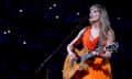 Taylor Swift iin an orange dress, playing a guitar and singing into a mic.