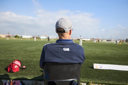 An older white man sits by a cricket pitch with his back to the camera, watching a game.