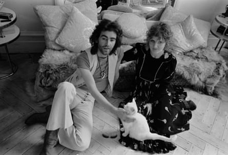 Black and white photo of Ossie Clark and Celia Birtwell taken in 1971, sitting on the floor in front of a sofa, with Ossie petting a cat that’s sitting on Celia’s dress, which is spread out on the floor in front of her