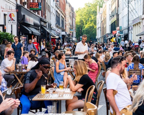 A crowded pedestrianised Soho street lined with outdoor diners at wooden tables, with shops and trees visible