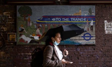 A woman with an umbrella walking past a mosaic sign saying "to the trains"