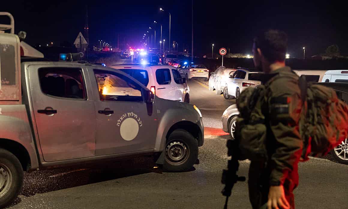 An Israeli soldier holding a gun stands next to vehicles on a road at night-time