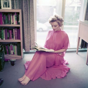 Marilyn Monroe in a flowing pink gown sits barefoot on the floor, reading a book