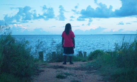 A scene from Descendant, with a person in a red shirt and black shorts gazing out at a water body