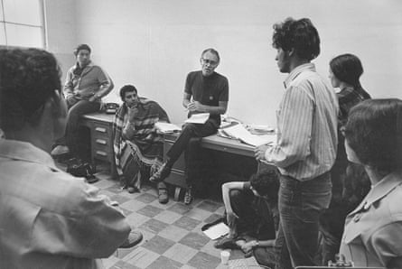 A man sitting on a table, surrounded by students standing and leaning against other tables.