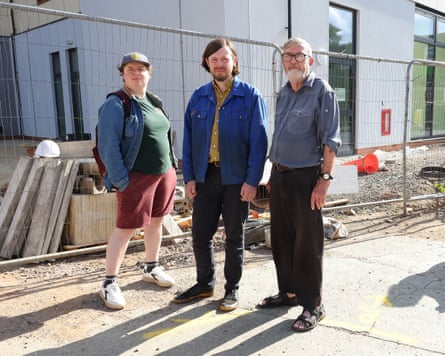 Three people lined up outside a fenced-off building site on a sunny day