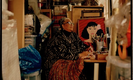 Rita Keegan sitting in a studio with piles of paintings and other materials, inclusdng one portrait of a young woman facing the camera