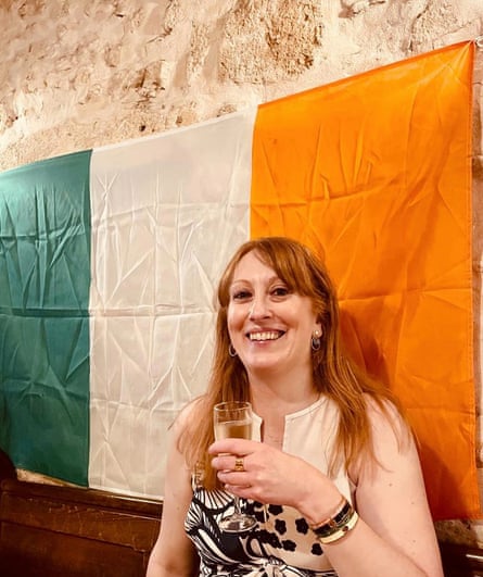 Karen Sugden holding a drink while sitting against an Irish flag on the wall behind her.