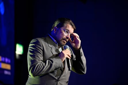 Jimmie Åkesson standing on stage at a political event holding a microphone and adjusting his glasses