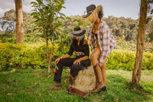 Two women, one standing adjusting her boot and the other sat on a hay bale while holding a red rose, both wear tassel-fringed cowboy/girl hats with tassel