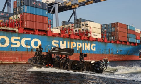 A black boat speeds through water next to a container ship