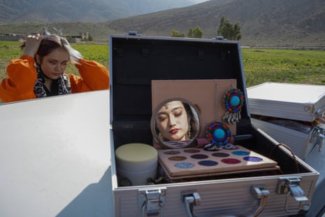 A woman looks into a mirror propped up in a makeup box, while opposite another woman ties up her hair.