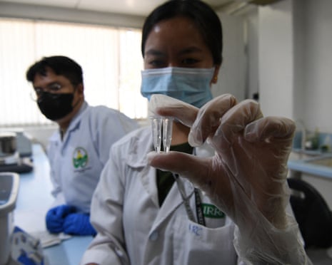 A woman wearing a face mask and a white laboratory coat holds up two pipettes as another researcher looks on