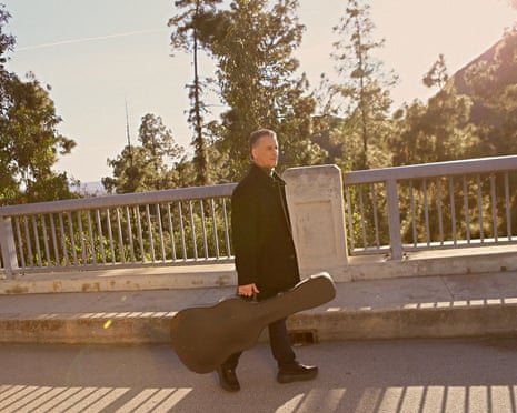 Daniel Levitin, carrying a guitar case, walks along a road with shadows cast by bright sun through railings behind him.