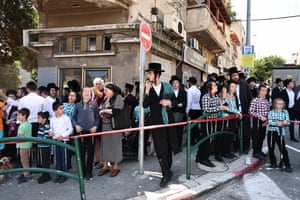 Men, women and children stand on a street corner behind a taped-off area, looking over at bomb damage that lies out of shot