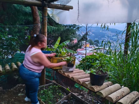 A woman waters plants on a balcony overlooking houses on a steep hillside.