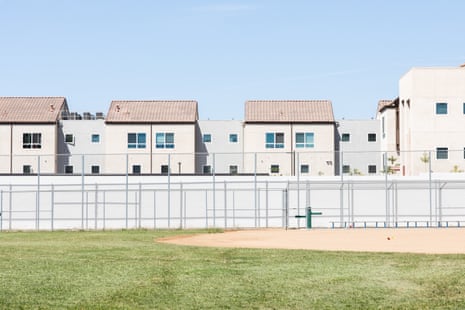 A row of light-colored buildings can be seen in back of a fenced softball field.