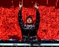 Kettama raises both arms above a DJ deck on stage, red lights behind