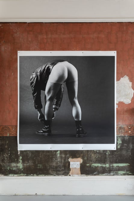 A photo of a man facing away from the camera bending over to tie his shoe laces. He is wearing a leather jacket, white underwear, no trousers and black leather boots.