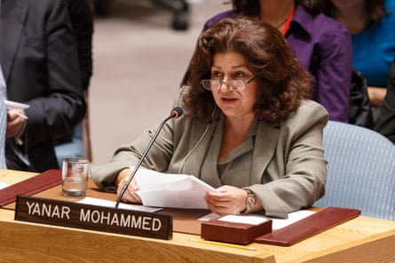 A middle-aged Arab woman reading from notes and speaking into a mic while sitting at a desk with her name on a sign