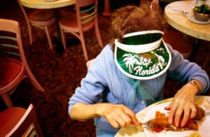 an older woman in a blue cardigan and a green sun visor hat obscuring her face sits at a table in a restaurant eating