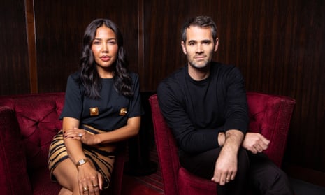 Emma and Jens Grede, both dressed in smart dark clothing, pose for a moodily lit portrait serated on red velvet armchairs in a wood-panelled room