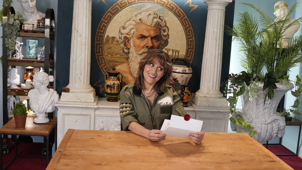 Nina Conti smiles while holding a sealed envelope at a wooden table