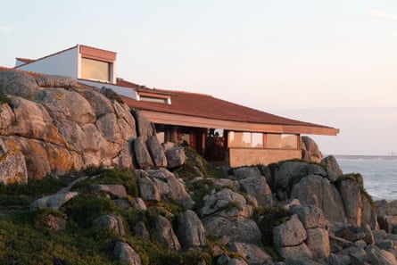 A shallow modernist building stands on rocks overlooking the sea, lit up by sunset.