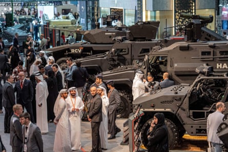 Visitors wearing traditional white Emirati robes and headdresses, with others in suits, walk along a row of military vehicles in an exhibition hall.