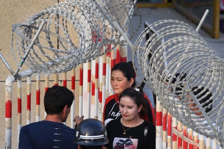 Two women walk between fences topped with razor wire, watched by a guard.
