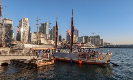 The double-hulled and masted canoe at a jetty with skyscrapers in the background
