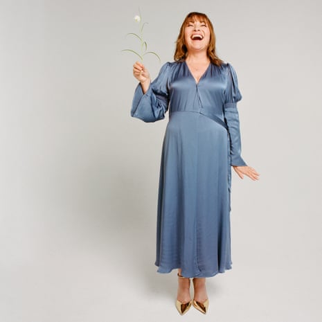 Lorraine Kelly standing holding a flower with her head back laughing, wearing a blue wrap dress and gold heels