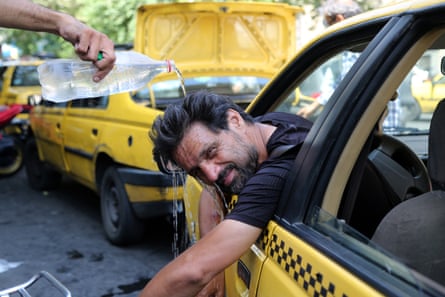 A taxi driver leans out of his car window and looks at the camera as a hand pours a bottle of water on to his head