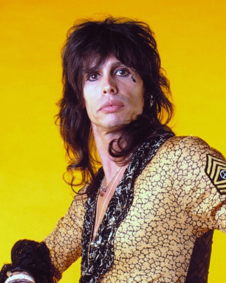 Tyler with a brown mullet that’s big on top and curly at the bottom, plus open-neck yellow leotard and lots of eyeliner