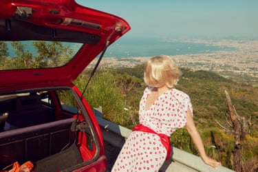 A woman in a polka dot dress sits on a low wall beside a red car overlooking Naples