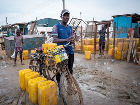 A man stands next to a bicycle and several yellow jerrycans. More jerrycans and two people can be seen behind him
