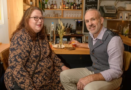 A woman with long red wavy hair, glasses and red lipstick, wearing a brown and black patterned dress, and a man with short grey hair, wearing a lilac shirt, a grey herringbone waistcoat and beige chinos, sitting at a restaurant table talking