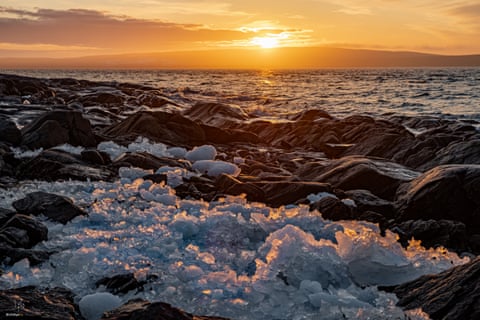The sun setting over the sea, with broken-up ice along a rocky shoreline in the foreground