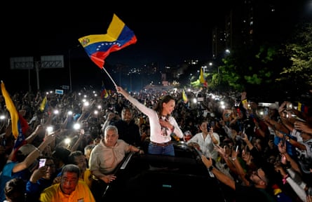 Maria Corina Machado stands through a car’s roof waving a flag above a cheering crowd