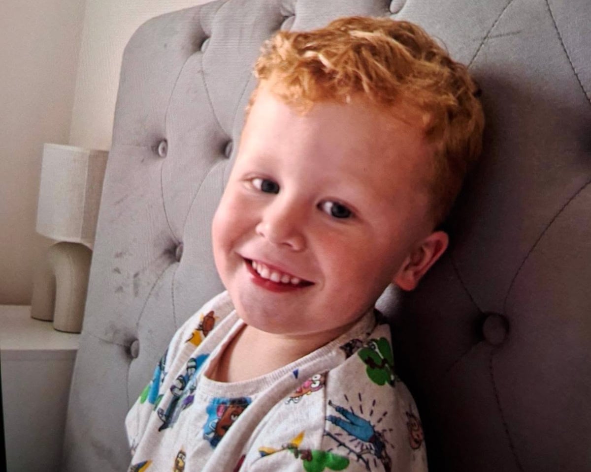 Noah Woods, who has curly ginger hair, is sitting against a headboard on a bed and smiling at the camera
