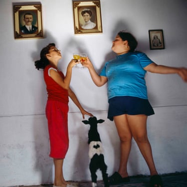 Two girls toast with drinks as a small goat watches, below framed portraits