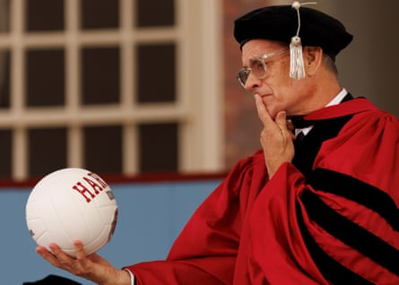 Tom Hanks wears red academic regalia as he gazes at a Harvard University emblazoned volleyball.