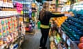 Seen from behind, a white woman with long blond hair reaches out a hand to select an avocado at a grocery store.