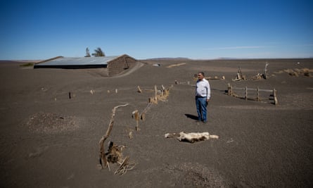 A man stands on loose brown earth in which fenceposts are submerged. Behind him a house is almost buried in the loose earth.